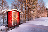 View of a rustic red wooden outhouse stands nestled amidst a serene, snow-covered landscape, birch trees dusted with frost, creating a tranquil winter scene, Finland.
