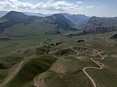 Luftaufnahme von kurvenreichen Straßen, die sich durch die grünen, sanften Hügel unter hoch aufragenden Bergen schlängeln, eine Symphonie aus Grün und Braun unter einem weiten Himmel, Naryn, Region Naryn, Kirgisistan.