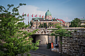 View of the Buda Castle, with its ornate facade and flags fluttering, framed by lush greenery and a stone bridge, Budapest, Central Hungary, Hungary.