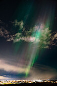 View of ethereal aurora borealis paints the night sky with strokes of green and purple above the distant city lights, Reykjavík, Reykjavíkurborg, Iceland.