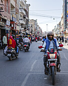 Bikaner, India - 24 June 2025: View of a bustling street scene, showcasing the vibrant life with scooters and pedestrians amidst aged buildings under a clear sky.