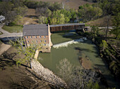 Aerial view of a historic mill clinging to the riverbank, its wooden bridge casting shadows on the rippling water, a rustic scene of nature and architecture, Missouri, United States.