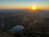 Luftaufnahme eines ruhigen Sees inmitten eines ausgedehnten Waldes, während die Sonne unter den Horizont sinkt und den Himmel in leuchtende Orange- und Goldtöne taucht, Feldberg, Baden-Württemberg, Deutschland.