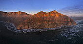 Luftaufnahme der zerklüfteten Küstenlinie, wo die goldenen Klippen auf den dunklen, aufgewühlten Ozean treffen und einen faszinierenden Kontrast aus Licht und Schatten erzeugen, Kapstadt, Westkap, Südafrika.