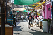 Bikaner, India - 24 June 2025: View of a cyclist weaving through bustling market street, vibrant awnings casting shadows on the road, framed by colorful buildings, creating a lively Indian scene.