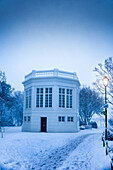 View of a snow-covered white building with a dark door and surrounding trees under a cold, blue-tinged sky, Reykjavík, Reykjavíkurborg, Iceland.