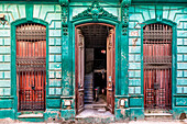 View of weathered turquoise building facade with ornate details frames open doors revealing a glimpse into life within, Havana, Havana, Cuba.