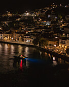 Aerial view of a town's waterfront illuminated by warm lights, reflected in the dark water where a boat cruises gently, Limni, Euboea, Greece.
