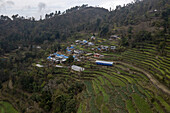 Luftaufnahme von terrassenförmig angelegten Feldern, die zu einem kleinen Dorf hinaufsteigen, das sich an die üppigen, zerklüfteten Berge schmiegt, ein Wandteppich aus Grün und Braun unter einem bewölkten Himmel, Jibjibe, Provinz Bagmati, Nepal.