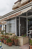 [Paphos, [Zypern] - 25. Juni 2025: Blick auf den 'KATASTROPAS SUPERMARKET' unter einer gestreiften Markise, umrahmt von Terrakotta-Töpfen mit Grünzeug, die einen Einblick in den lokalen zyprischen Handel bieten.
