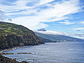 Luftaufnahme der zerklüfteten Küstenlinie, die auf den ruhigen Ozean trifft, mit dem Berg Pico, der in der Ferne die Wolken durchbricht, Vulkankrater Pico, Azoren, Portugal.