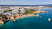 Aerial view of golden cliffs meet the turquoise sea, framing sandy beaches and modern buildings in a vibrant coastal scene, Lagos, Algarve, Portugal.