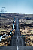 View of a long, stark road stretching through a barren landscape, juxtaposed with industrial pipelines and distant power lines, Nesjavellir, Grímsnes- og Grafningshreppur, Iceland.