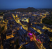 Aerial view of streets and buildings glow warmly against the twilight sky, Arthur's Seat looms in the distance, creating a dramatic contrast between urban illumination and natural darkness, Edinburgh, Scotland, United Kingdom.