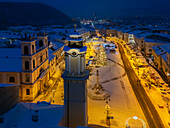 Aerial view of the snow-covered Námestie SNP square with illuminated buildings, a towering column, and Christmas trees casting a warm glow, Banská Bystrica, Banskobystrický kraj, Slovakia.
