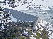 Aerial view of the imposing concrete dam rises amidst the snowy landscape and icy waters, contrasted by a small red building and sparse trees, Saint-Maurice, Valais, Switzerland.