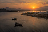 Blick auf das ruhige Wasser, in dem sich die untergehende Sonne spiegelt und die Berge und Boote in der Ferne in ein goldenes Licht taucht - eine friedliche Szene, Sur, Oman.