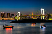 View of illuminated boats sailing on dark waters beneath a brightly lit bridge and city skyline under a twilight sky, Tokyo, Tokyo, Japan.