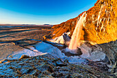 View of golden sunlight illuminates the cascading Seljalandsfoss waterfall against the stark Icelandic landscape, with snow-dusted ground, , Iceland.