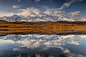 View of snow-capped mountains mirrored in the still lake waters under a sky brushed with clouds, a serene landscape of cold peaks and golden trees, Altai Republic, Russia.