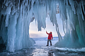 Baikal, Russia - 27 January 2022: View of a figure in red standing beneath a frozen grotto of shimmering icicles, reaching out towards the icy formations, against the backdrop of a winter landscape.