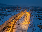 Aerial view of snow-covered Námestie SNP, illuminated by warm lights contrasting with the cold blue dusk, Banská Bystrica, Banskobystrický kraj, Slovakia.
