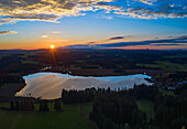 Luftaufnahme des ruhigen Sees, in dem sich der leuchtende Sonnenuntergang spiegelt, eingerahmt von dunklen Wäldern und offenen Feldern, die eine ruhige Szene schaffen, Aitrang, Bayern, Deutschland.