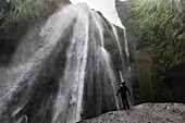 Blick auf einen mächtigen Wasserfall, der über moosbewachsene Klippen stürzt. Eine einsame Gestalt steht in Ehrfurcht vor der rauen Schönheit der Natur und spürt den Nebel, Island.