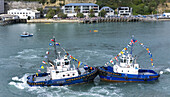 Nelson, New Zealand - 18 June 2025: Aerial view of tugboats adorned with vibrant flags create swirling patterns on the water near the harbor, framed by buildings and a pier.