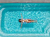 Aerial view of a woman in a white bikini floats serenely on a purple inflatable mattress, embraced by the crystalline turquoise waters of a swimming pool, Strasbourg, Grand Est, France.