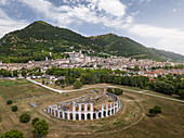 Aerial view of the Roman Theatre's ancient stones contrast with the modern city nestled against the verdant hills, Gubbio, Perugia Province, Italy.