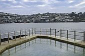Blick auf das ruhige Wasser, in dem sich die Gebäude von Polruan bei bewölktem Himmel spiegeln, eingerahmt von der Struktur des White House Sea Pools mit Metallgeländer, Fowey, England, Vereinigtes Königreich.