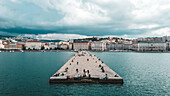 Luftaufnahme eines Piers, der sich in die Adria erstreckt, und eines belebten Weges vor dem Hintergrund der Skyline der Stadt Triest, Friaul-Julisch Venetien, Italien.