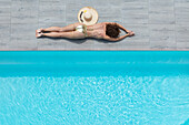 Aerial view of a sunbather with a straw hat on grey tiles beside the clear turquoise waters of a swimming pool, Strasbourg, Grand Est, France.