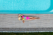 Aerial view of a woman sunbathing by a pool with crystal-clear water, a vibrant pink and yellow swimsuit contrasting against the cool gray tiles, Strasbourg, Grand Est, France.
