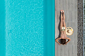 Aerial view of a woman relaxing beside a turquoise pool, her straw hat casting a shadow on the warm grey tiles, creating a tranquil summer scene, Strasbourg, Grand Est, France.