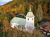 Aerial view of a church with a green roof standing prominently against a backdrop of autumn-colored trees, bathed in the soft light, Blatnica, Žilina Region, Slovakia.