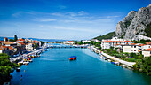 Luftaufnahme des Flusses Cetina, der in die Adria mündet, mit an der Uferpromenade angedockten Booten und dem Dinara-Gebirge im Hintergrund, Omiš, Kroatien.
