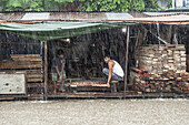 Chattogram, Bangladesh - 05 August 2023: View of two men working with wood under a makeshift shelter during a heavy downpour, rain blurring the scene into a hazy, wet tableau.