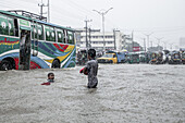 Chattogram, Bangladesch - 05. August 2023: Blick auf Kinder, die durch die Fluten einer Stadtstraße waten, in der bunte Busse und Fahrzeuge teilweise überflutet sind.