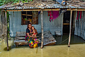 Bogura, Bangladesch - 07. März 2020: Blick auf eine Mutter, die ihr Kind auf einer hölzernen Plattform inmitten der Überschwemmungen hält. Das Wellblechhaus steht in starkem Kontrast zum trüben Wasser.