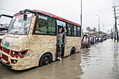 Chattogram, Bangladesh - 05 August 2023: View of a bus passenger standing by the door in floodwater, with rickshaws lined up along the submerged road, reflecting the overcast sky.