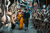 Kolkata, India - 28 January 2023: View of a woman in a vibrant yellow sari and a boy in school uniform walking amidst the clay idols of Kumartuli.