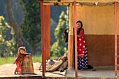 Dhangadhi, Nepal - 03 March 2023: View of a woman standing near a rustic building with red pillars, while a young girl plays with a basket, bathed in warm sunlight.