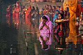 Bogura, Bangladesh - 20 November 2023: View of women in colorful saris standing in tranquil waters, holding offerings, their reflections shimmering against the dark river surface.