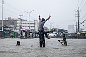 Chattogram, Bangladesch - 05. August 2023: Blick auf Kinder, die fröhlich durch die überfluteten Straßen in der Nähe von Agrabad planschen. Ihr Lachen hallt inmitten des grauen Himmels und der vom Regen durchnässten Gebäude wider.
