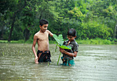 Mirsarai, Bangladesch - 01. Januar 2000: Blick auf zwei Jungen, die durch die überschwemmten Felder waten. Der eine hält ein großes grünes Blatt in der Hand, während der Regen fällt und die Grüntöne der umliegenden Bäume widerspiegelt.