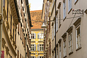 View of narrow alleyway with buildings on both sides, light suspended overhead, and a glimpse of yellow facade in the distance, Graz, Austria.