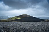 View of a solitary, dark volcanic cone rises starkly from a vast, grey gravel plain under a brooding sky, a scene of stark beauty, Kirkjubaejarklaustur, Skaftárhreppur, Iceland.