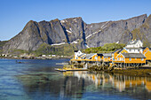 View of vibrant yellow and white houses perched on stilts reflect in the tranquil blue waters beneath the towering, snow-dusted mountains, Hamnoy, Nordland, Norway.